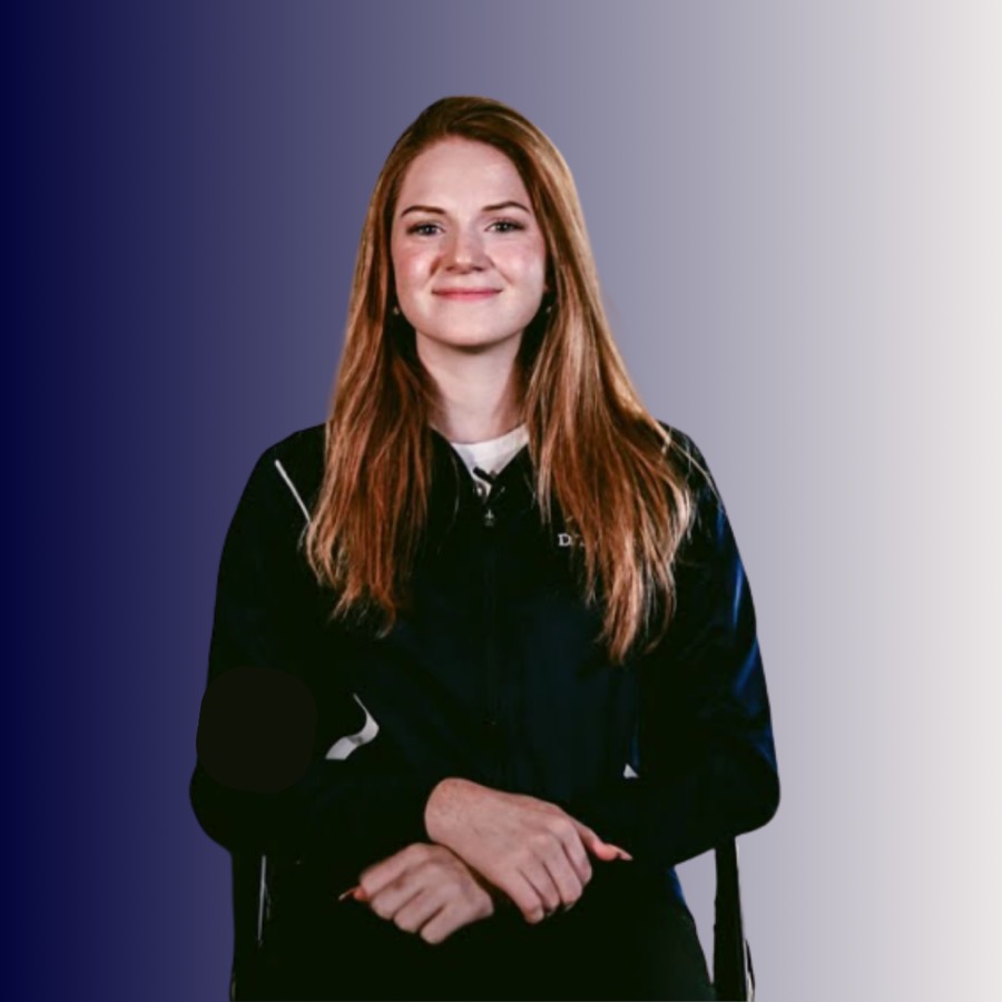 A young woman with long red hair, wearing a black jacket, standing with her arms crossed against a gradient purple background.